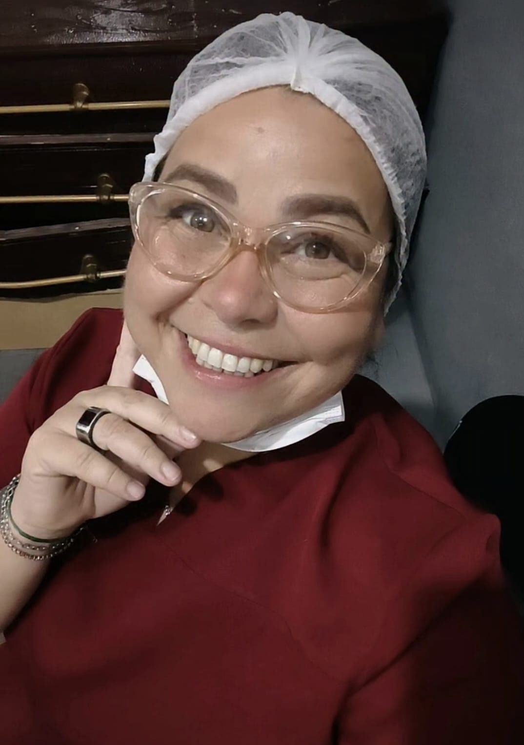 foto de uma mulher sorrindo, usando óculos claros, touca branca e camisa vermelha, com roupas de laboratório n
